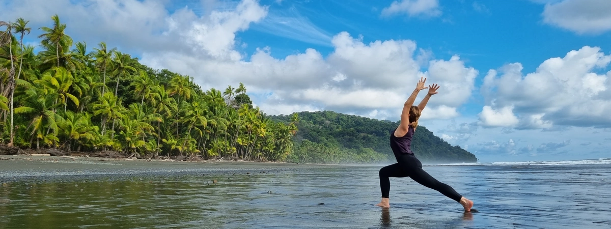Claudia Schriever steht am Strand mit Blick auf Palmen gerichtet in Yogakleidung in der Yoga Position High Lunge