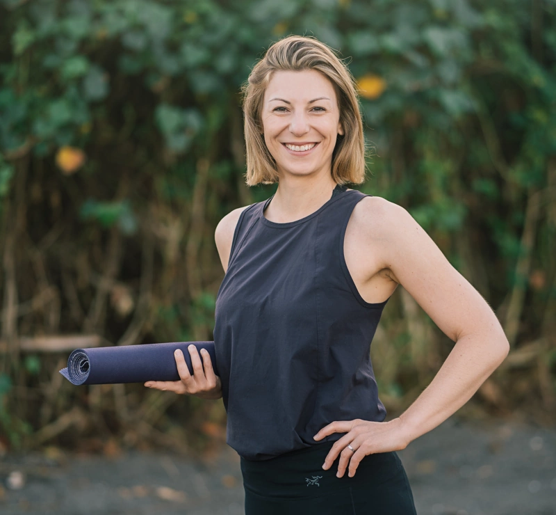 Profilfoto von Claudia Schriever in Yogagewand mit zusammengerollter Yogamatte in einer Hand